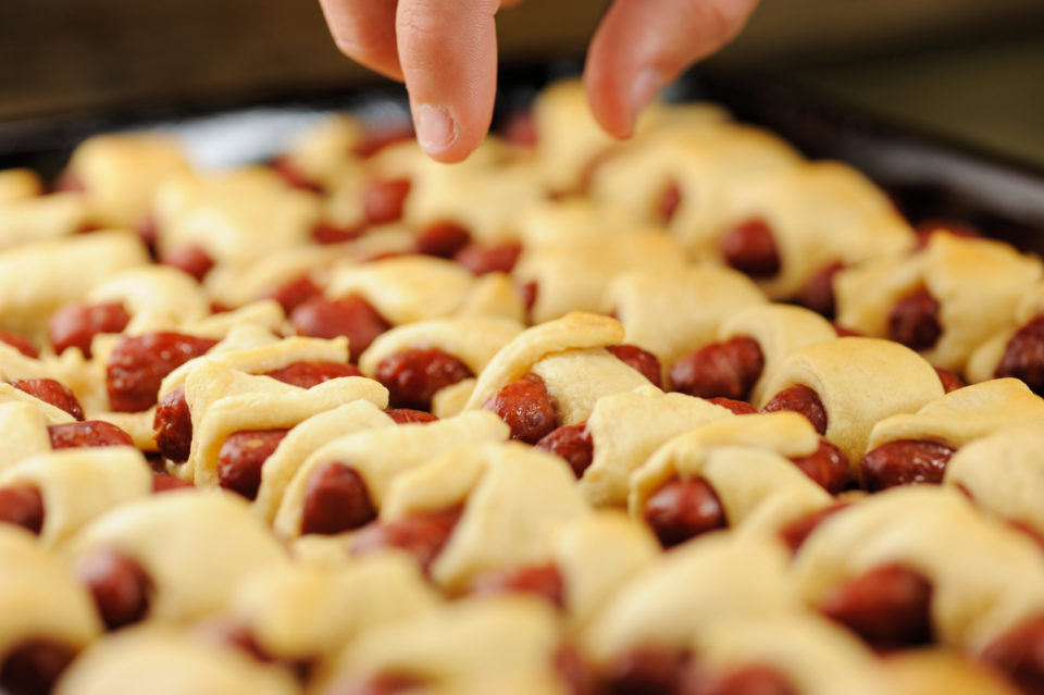 Hand selecting pigs in a blanket appetizer sausage roll from kitchen tray after being cooked.