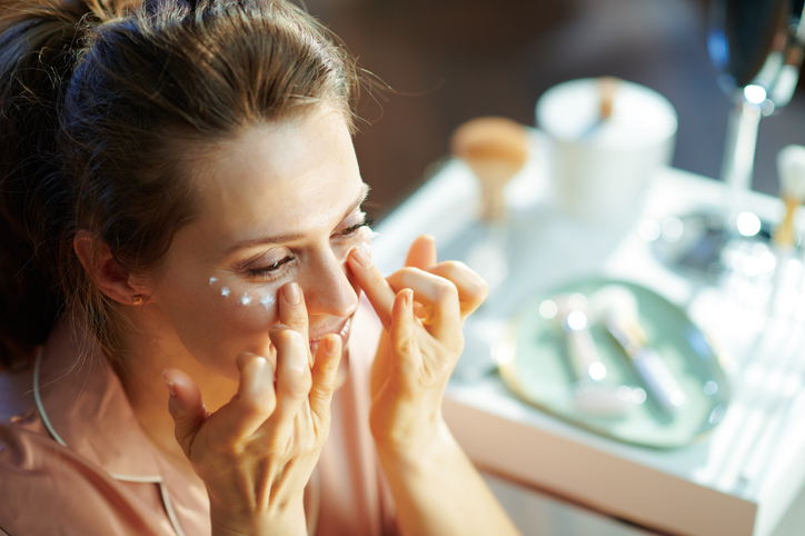 elegant female in pajamas at modern home in sunny winter day applying eye cream.