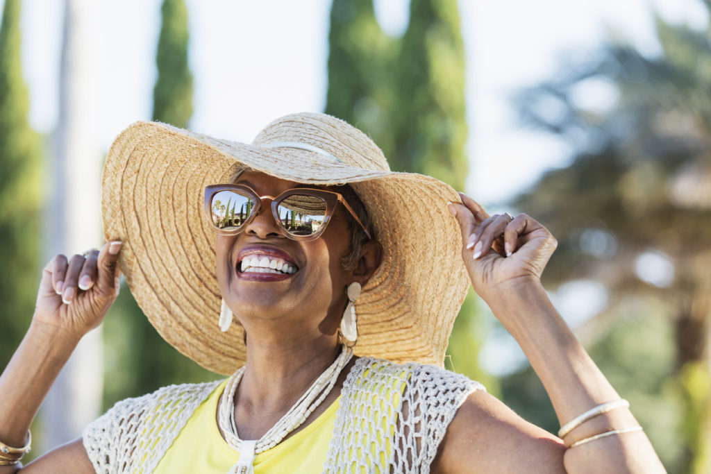 African-American woman wearing sunglasses and a wide brim hat on a sunny day. She is looking up at the sky, smiling. A building, palm trees and clear blue skies are visible in the reflection in the sunglasses.